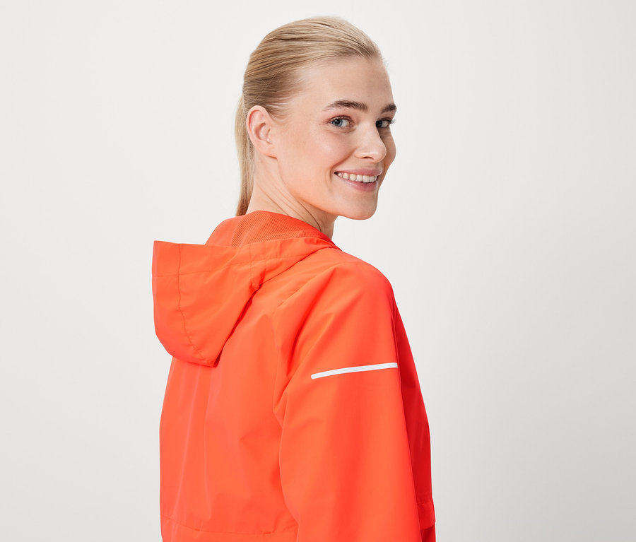 Eine Frau lächelt über die Schulter und trägt eine orangefarbene Jacke mit Kapuze und reflektierendem Streifen.