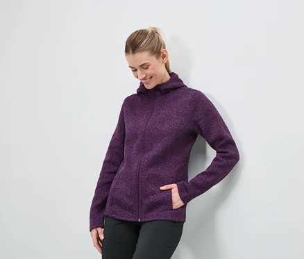 Eine Frau in einer lila Kapuzenjacke mit Reißverschluss und einer schwarzen Hose steht an einer weißen Wand.