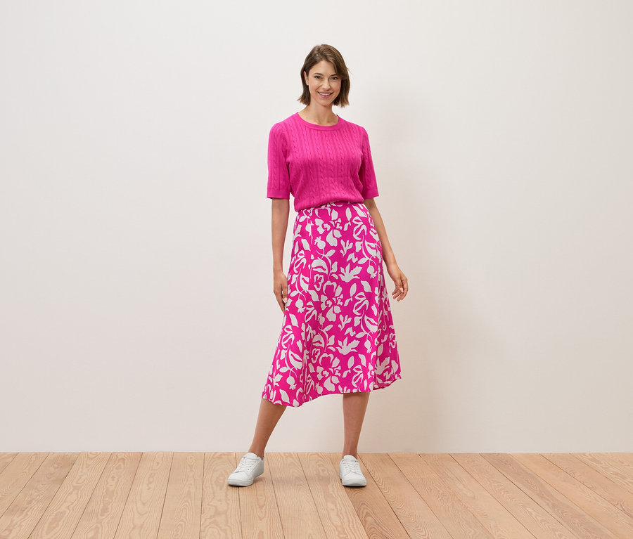Frau steht, trägt ein pinkes Oberteil und einen Rock mit Blumenmuster.