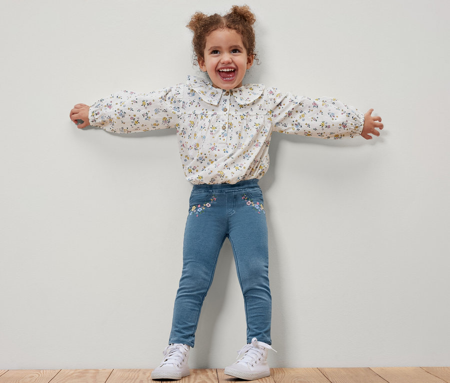 Ein lächelndes Mädchen steht mit ausgestreckten Armen und trägt Kinder-Jeggings – Fit »Mia« und eine geblümte Bluse.