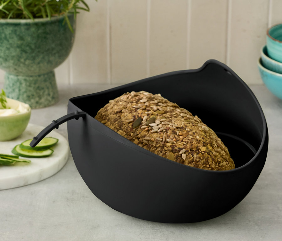 Vollkornbrot in schwarzem Behälter, daneben Butter, Gurke und eine Topfpflanze.