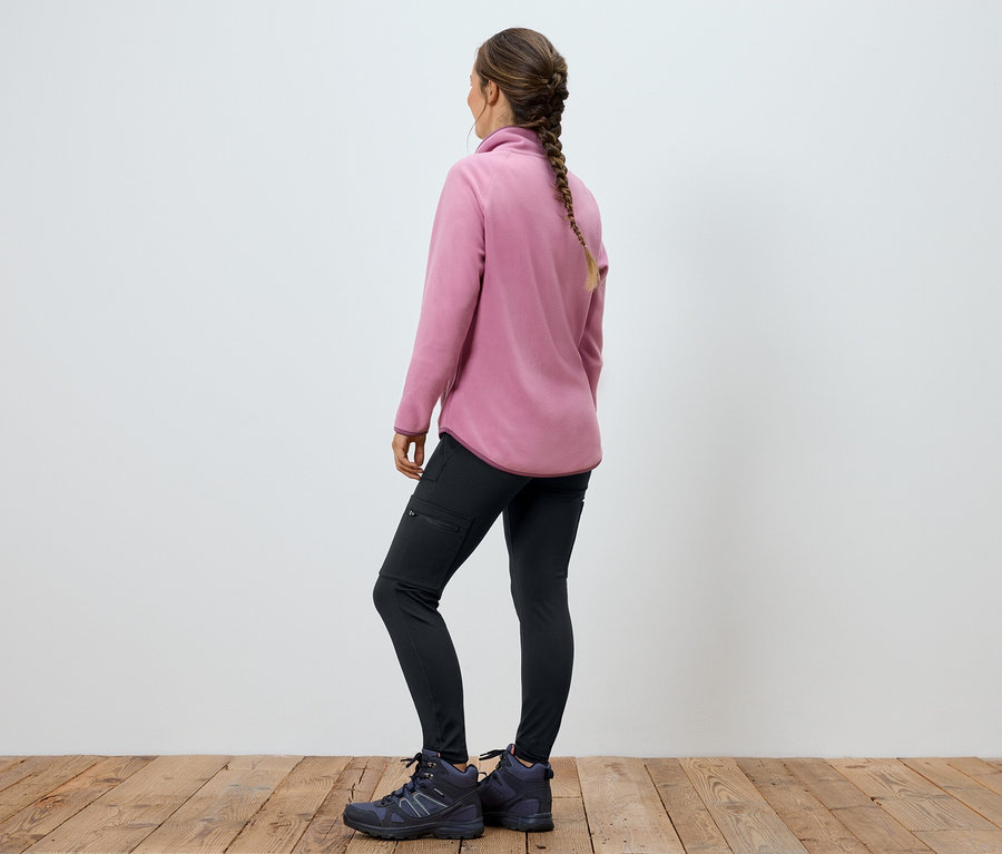 Frau in rosa Fleecejacke, schwarzen Hosen und Stiefeln steht mit dem Rücken zur Kamera.