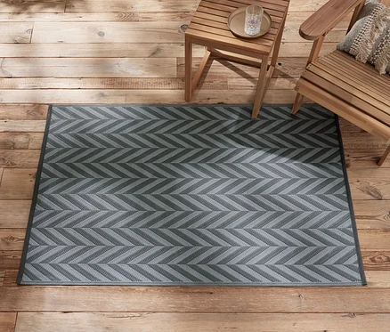 Ein ca. 165 x 115 cm großer, anthrazit-hellgrauer Outdoor-Teppich liegt auf einem Holzboden neben hölzernen Gartenmöbeln.