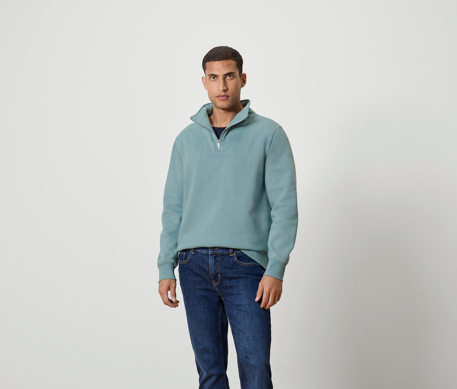 Mann steht und trägt ein blaugrünes Sweatshirt mit Reißverschluss und Jeans.