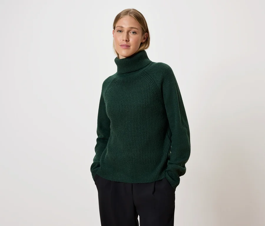 Frau posiert in einem grünen Rollkragenpullover und einer schwarzen Hose.