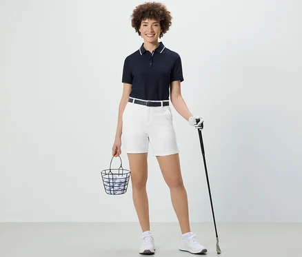 Frau hält einen Korb mit Golfbällen und einen Golfschläger. Sie trägt weiße Softshell-Funktionsshorts.