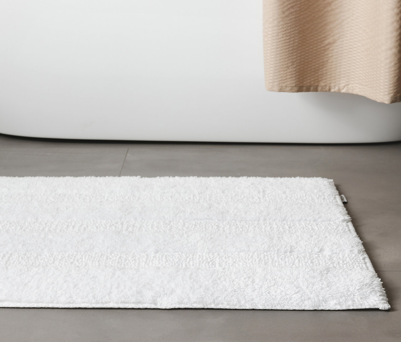 Weiße, rechteckige Badematte »Ilka« von benuta (50 x 80 cm) liegt auf grauem Fliesenboden vor einer Badewanne.