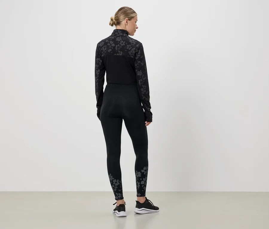 Rückansicht einer Frau mit Pferdeschwanz in schwarzer Sportbekleidung mit Blumenmuster und schwarzen Turnschuhen vor einer weißen Wand.
