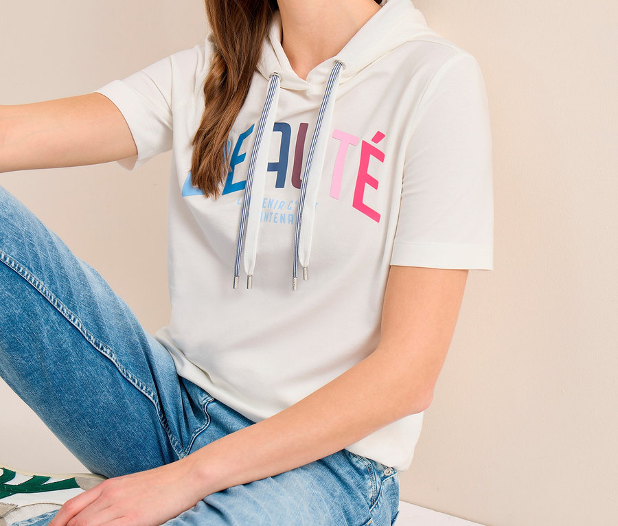 Frau sitzt am Boden und trägt ein weißes kurzärmeliges CECIL Hoodie-Shirt mit buntem Aufdruck, hellblaue Jeans und weiße Sneaker.