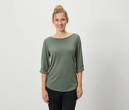 Eine Frau mit hochgesteckten blonden Haaren trägt ein olivgrünes Langarmshirt und schwarze Hosen vor einem weißen Hintergrund.