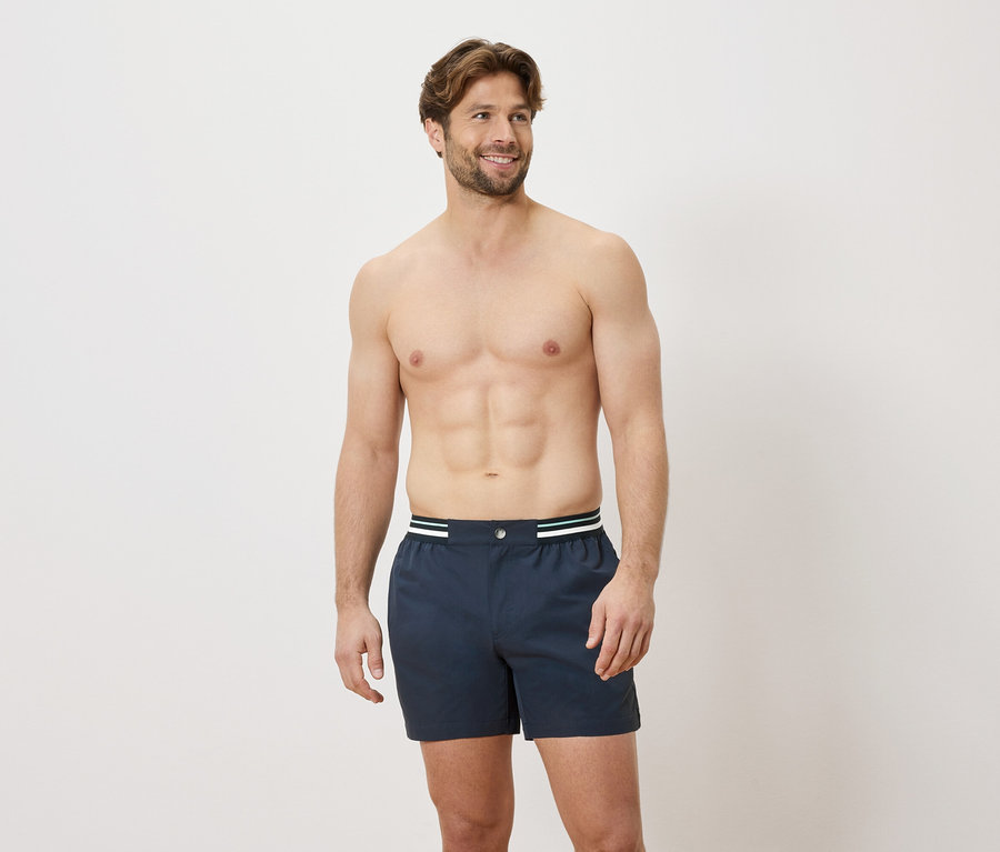 Ein Mann mit freiem Oberkörper trägt blaue Boxershorts mit einem weißen und grünen Bund vor einem weißen Hintergrund.