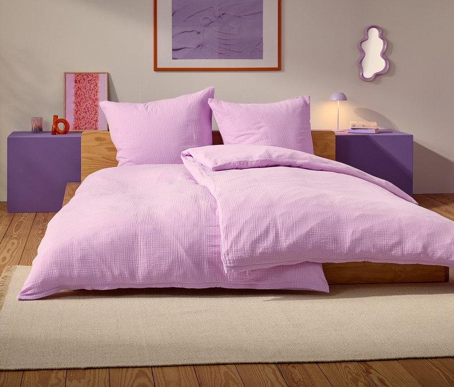 Ein lila Bett mit zwei Kissen und einer Decke steht auf einem beige Teppich in einem Schlafzimmer.