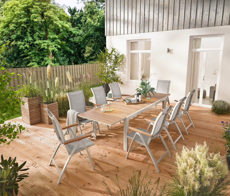 Ein ausziehbarer Gartentisch aus Aluminium »Liska« mit ca. 200–300 cm Länge steht auf einer Holzterrasse, umgeben von sechs Hochlehnern »Liska« mit verstellbarer Rückenlehne. Zwei Pflanzsäulen »Tinus« und fünf LED-Gartenfackeln sind ebenfalls sichtbar.