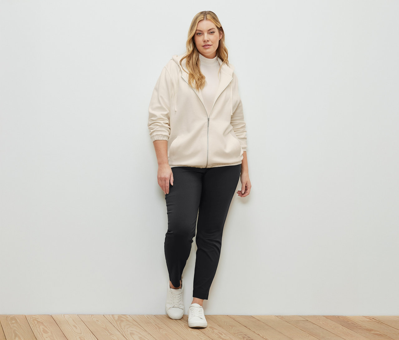 Frau steht und trägt eine cremeweiße Sweatjacke, ein cremeweißes Blusenshirt mit Stehkragen und eine Stretchhose.