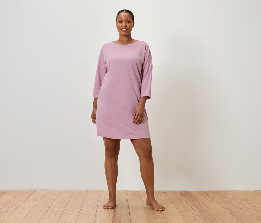 Eine Frau steht barfuß vor einer Wand und trägt ein rosafarbenes, kurzärmeliges Kleid.