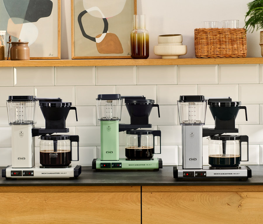 Drei Filterkaffeemaschinen »Moccamaster KBG Select« in Weiß, Pastellgrün und gebürstetem Aluminium stehen nebeneinander auf einer Arbeitsplatte. Jede Maschine hat eine Glaskanne mit Kaffee.