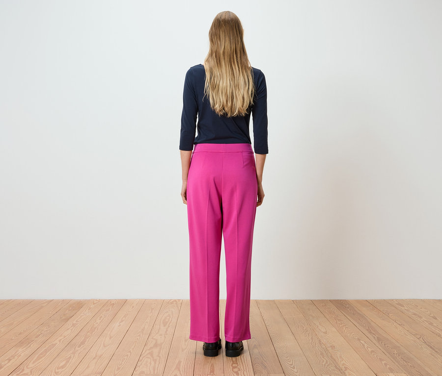 Rückansicht einer Frau mit langen blonden Haaren, die ein marineblaues Langarmshirt und eine pinke Hose trägt.