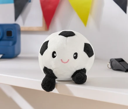 Ein weißer Stofffußball mit schwarzen Flecken, einem Gesicht und kleinen Beinen steht auf einer weißen Oberfläche, daneben eine blaue Kamera und eine schwarze Trillerpfeife.