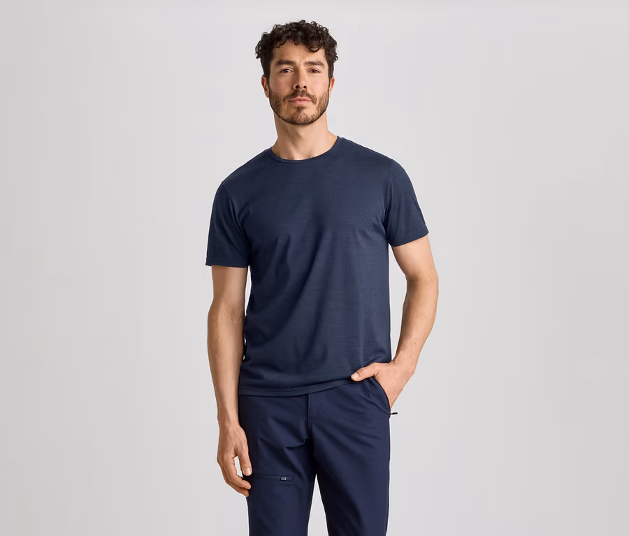 Ein Mann mit lockigem Haar und Bart trägt ein dunkelblaues T-Shirt und eine dunkelblaue Hose. Er steht mit der rechten Hand in der Hosentasche.