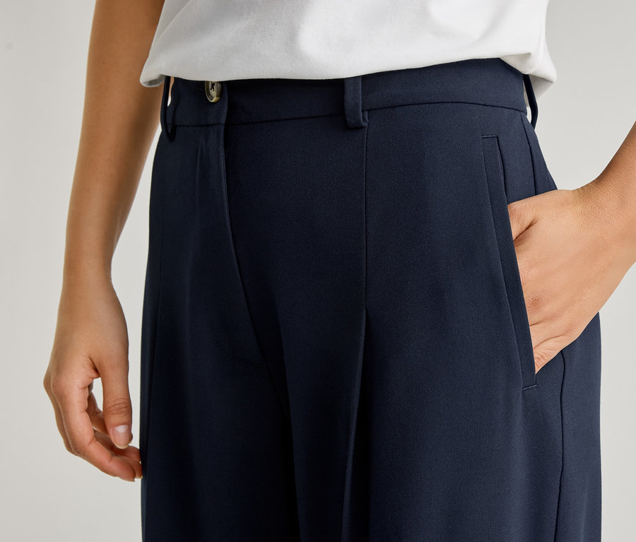 Nahaufnahme einer Person mit einer Hand in der Tasche einer marineblauen Hose mit Bundfalten und Knopf.