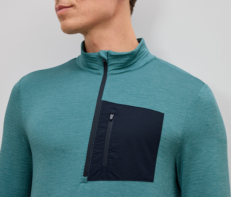 Nahaufnahme eines Mannes, der ein türkisfarbenes Langarmshirt mit kurzem Reißverschluss und einer einzelnen aufgesetzten Reißverschlusstasche auf der linken Seite trägt.