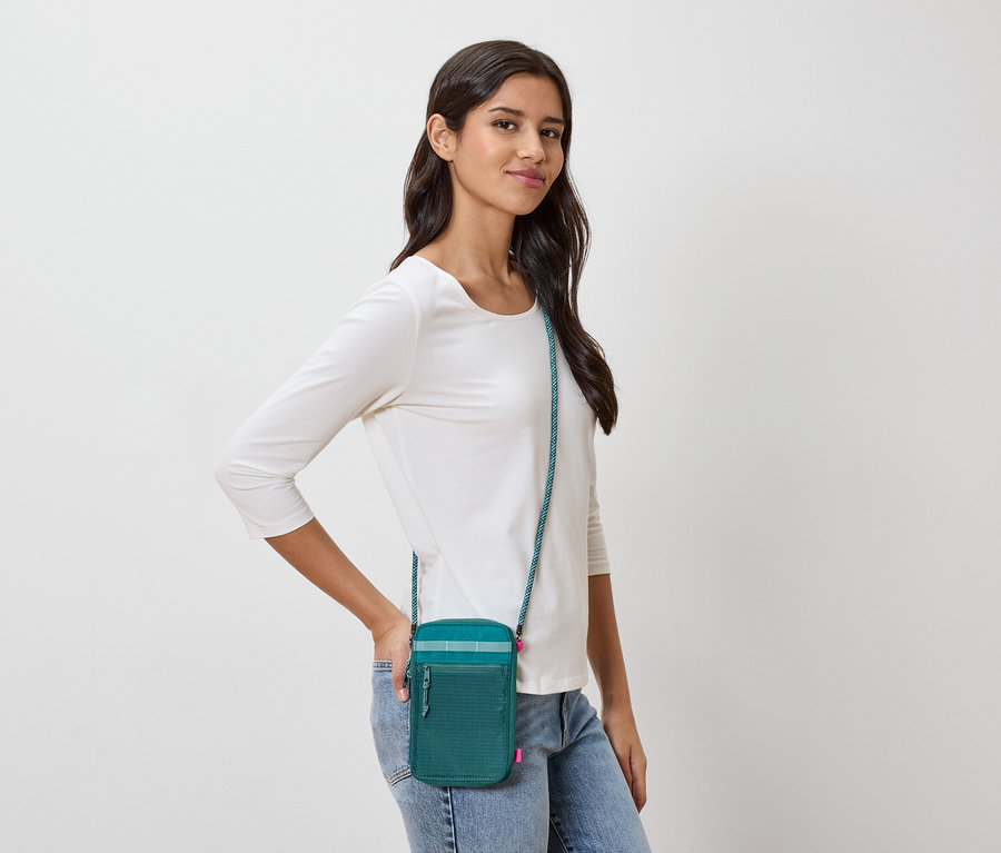 Frau mit dunkelbraunen Haaren, die eine grüne Umhängetasche mit blaugrünem Trageriemen und ein weißes Langarmshirt und Jeans trägt.