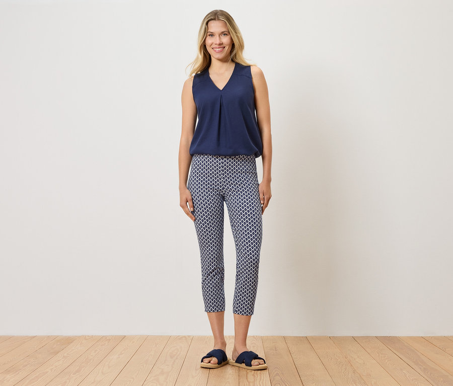 Frau steht und trägt ein blaues Top, gemusterte Hose und blaue Hausschuhe.