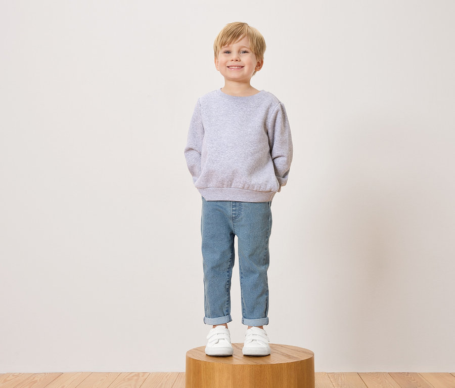 Ein lächelnder Junge mit blonden Haaren steht auf einem Holzhocker und trägt einen grauen Pullover, Jeans und weiße Turnschuhe.