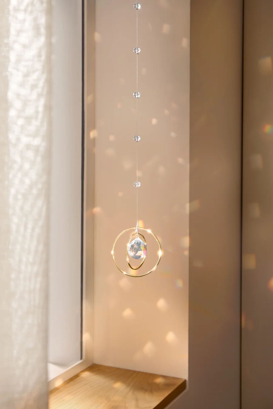 Ein Kristall-Mobile mit goldenen Ringen hängt vor einem Fenster und wirft Lichtflecken an die Wand.