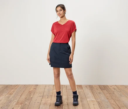 Frau steht vor einer weißen Wand, trägt ein rotes T-Shirt, einen blauen Rock und blaue Wanderschuhe.