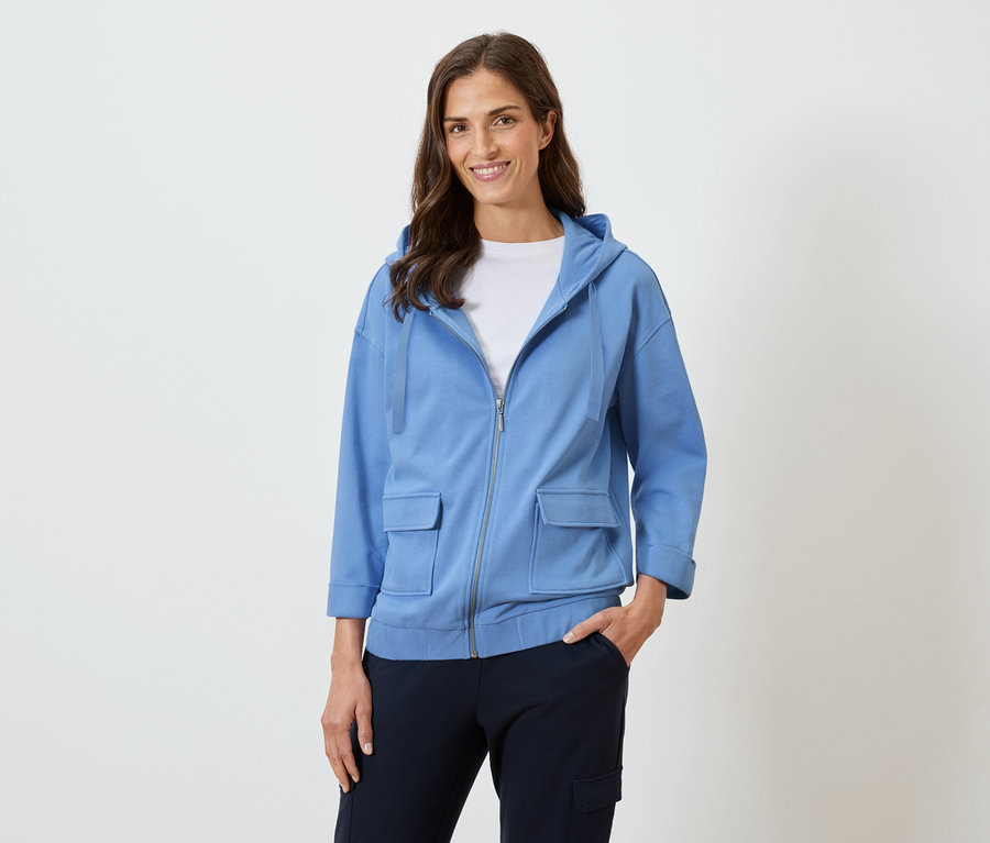 Frau mit langen braunen Haaren lächelt in einer blauen Kapuzenjacke und dunkler Hose.