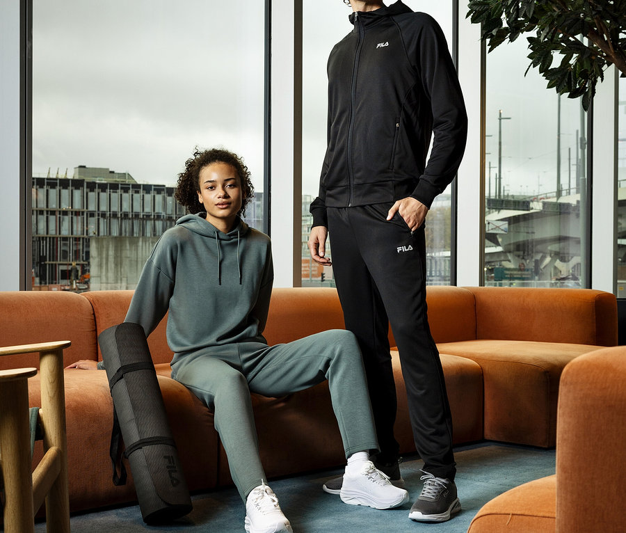 Eine junge Frau sitzt mit einer gerollten Yogamatte neben sich auf einem orangefarbenen Sofa, während ein junger Mann daneben steht. Beide tragen Trainingsanzüge und Turnschuhe.