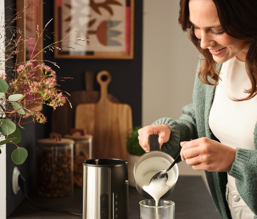 Frau gießt Milchschaum mit einem Löffel in ein Glas Kaffee neben einem Milchaufschäumer.