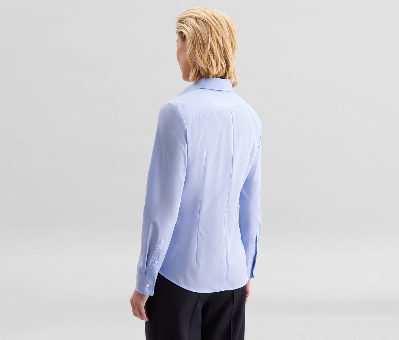 Frau von hinten, mit kurzen blonden Haaren, trägt eine hellblaue Seidensticker Bluse Slim Fit non iron und schwarze Hose.
