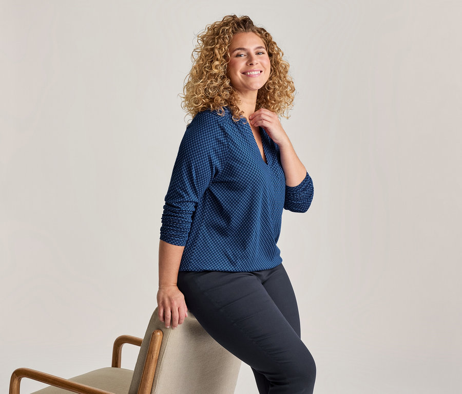 Lächelnde Frau mit lockigem blondem Haar, die eine blau gemusterte Bluse und dunkle Hose trägt und seitlich auf der Rückenlehne eines hellen Stuhls sitzt.