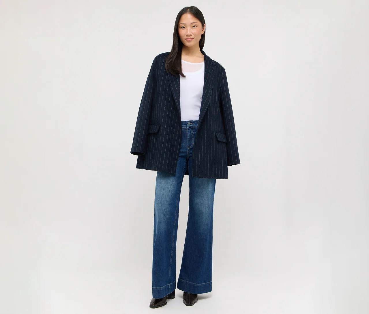 Eine Frau mit dunklen Haaren steht und trägt einen dunkelblauen Nadelstreifenblazer über einem weißen Oberteil, weite blaue Jeans und dunkle Schuhe.