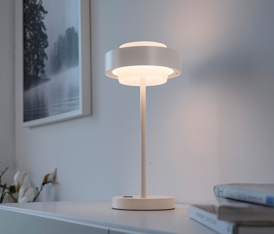 Weisse Tischlampe mit geschichtetem, rundem Schirm leuchtet auf einem weissen Schrank. Dahinter ein gerahmtes Bild einer nebligen Landschaft, rechts Bücherstapel, links weisse Blumen.