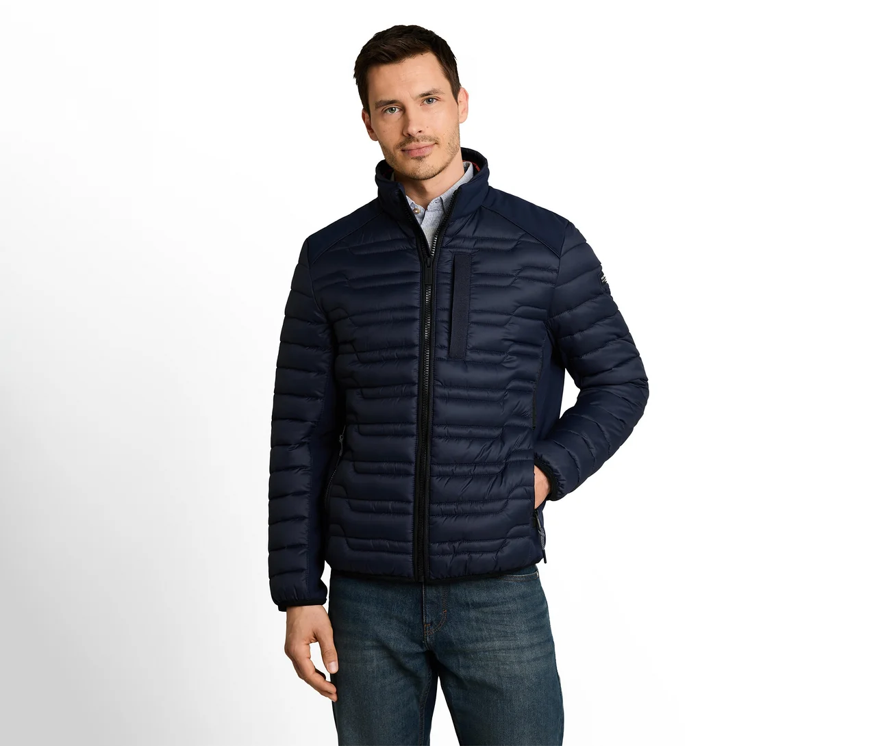 Mann trägt eine dunkelblaue Steppjacke und blaue Jeans, steht mit einer Hand in der Tasche und blickt nach vorne.