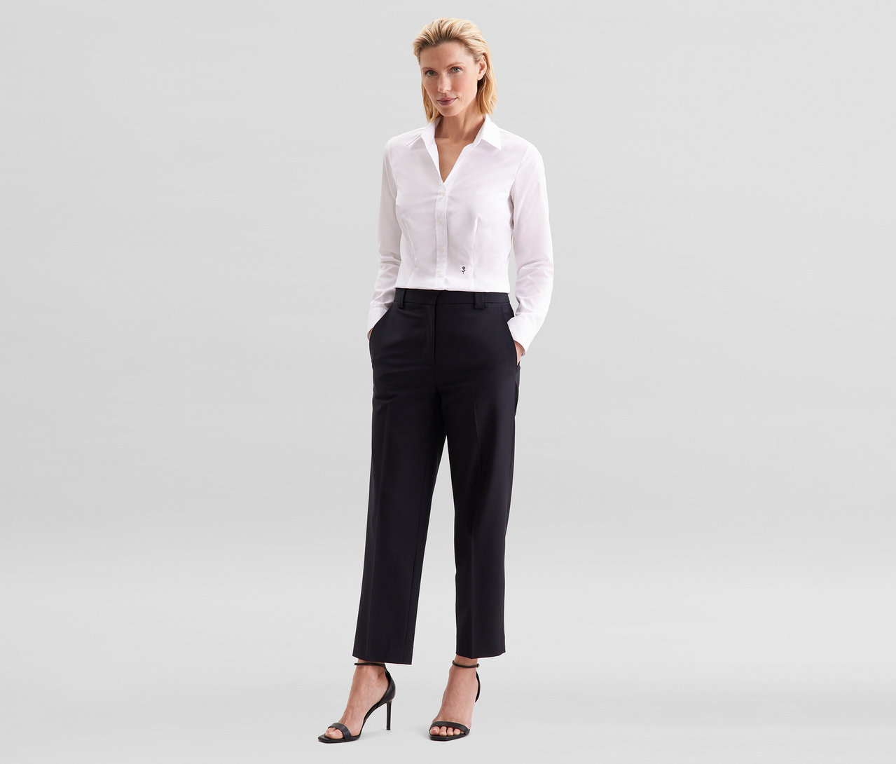 Eine Frau mit blonden Haaren steht mit den Händen in den Taschen ihrer schwarzen Hose. Sie trägt eine weiße Seidensticker Bluse Slim Fit non iron und schwarze Sandaletten mit hohen Absätzen.