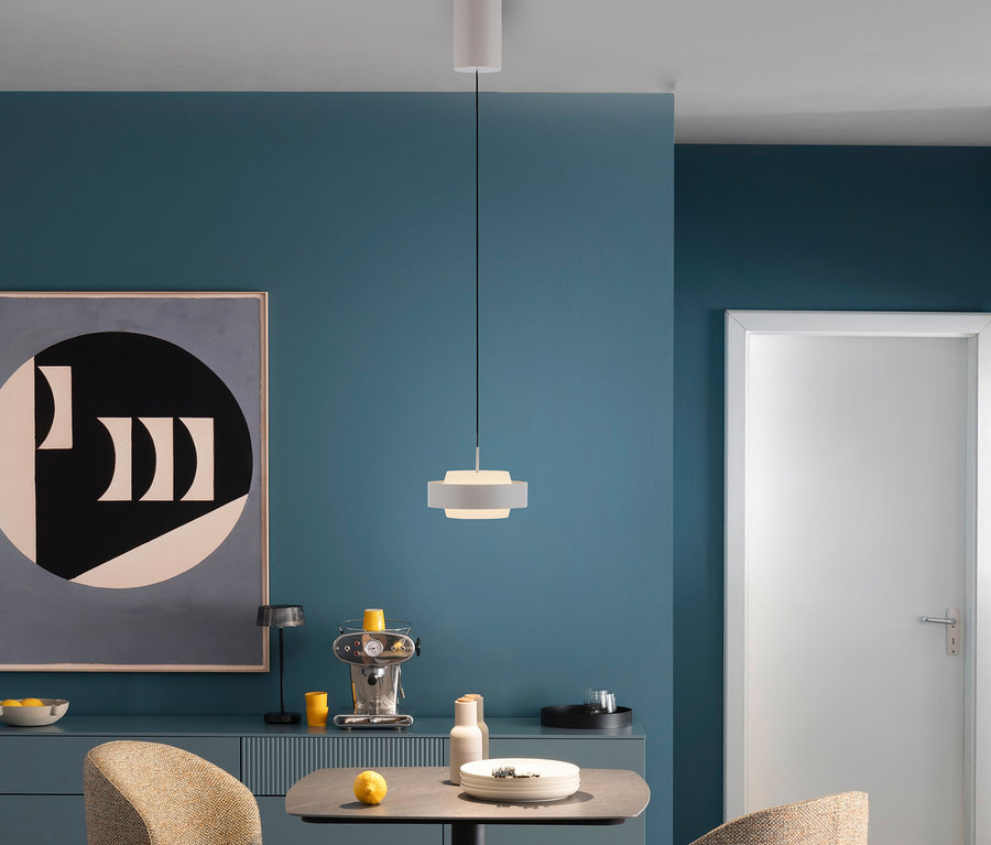 Ein modernes Esszimmer mit tealfarbenen Wänden, einer weißen Pendelleuchte über einem grauen Tisch mit Tellern und einer Zitrone. Links steht ein Sideboard mit einer Espressomaschine und abstrakter Kunst, rechts eine weiße Tür.