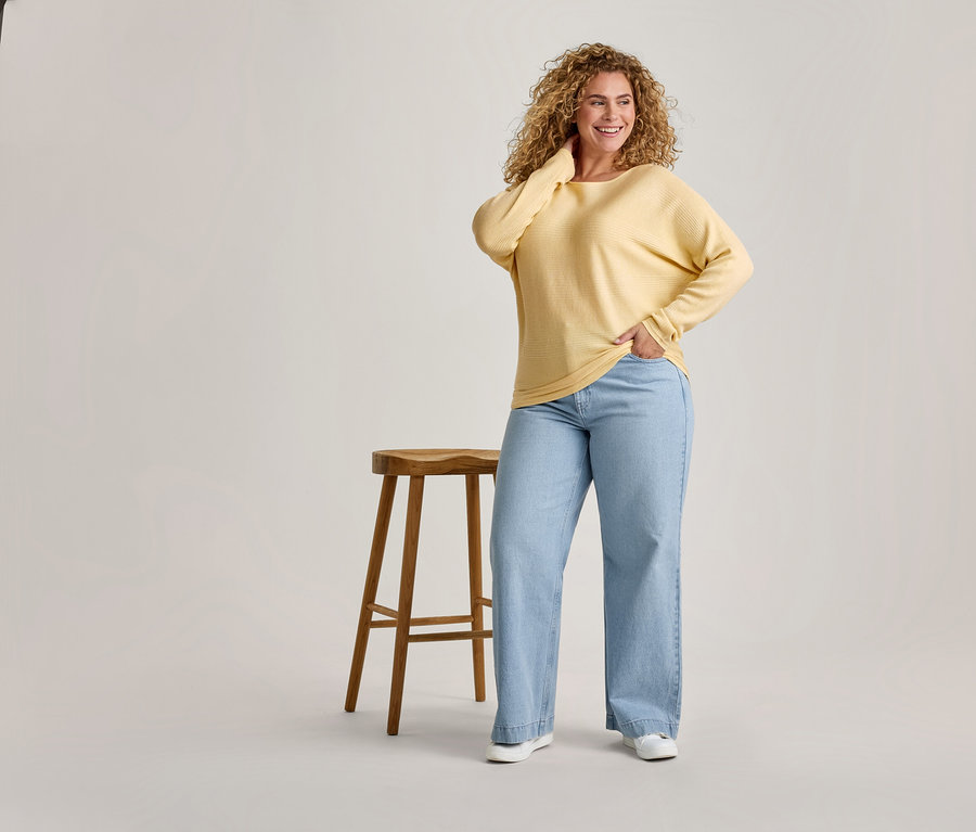Lächelnde Frau mit lockigem Haar, die ein gelbes Langarmshirt und hellblaue weite Jeans trägt, steht neben einem Holzschemel.