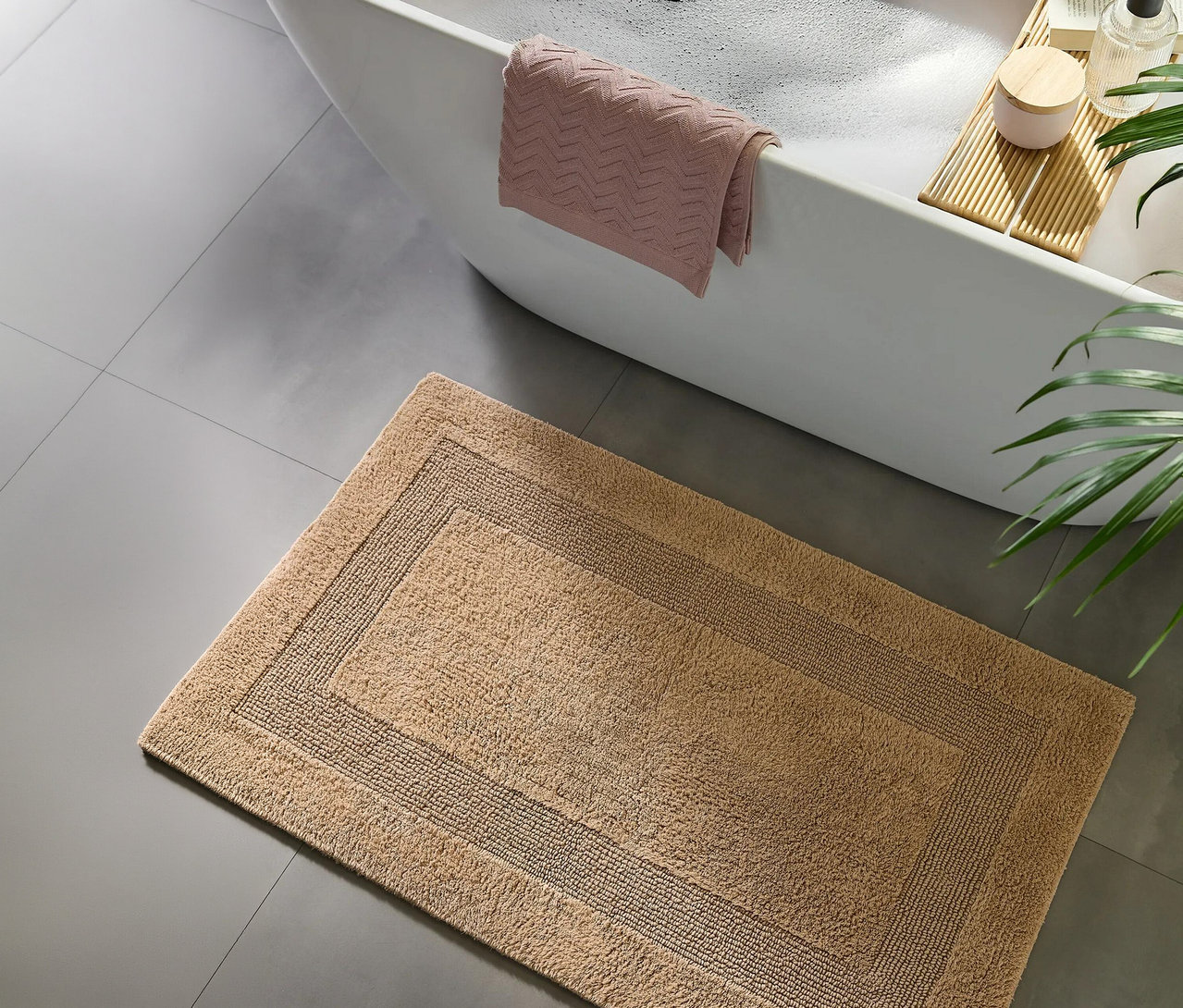 Eine hellbraune Badematte mit dunklerem Rand auf grauem Fliesenboden, vor einer weißen Badewanne mit rosa Handtuch und Holzwannenablage.