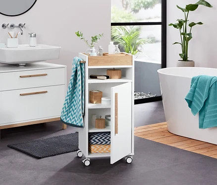 Im Badezimmer stehen ein weißer Waschbeckenunterschrank »Eklund« mit zwei Schubladen, ein Utensilo, ein Seifenspender, ein Frottier-Badetuch, 2 Jacquard-Handtücher, 2 Frottier-Handtücher (anthrazit), 2 Dosen aus Bambus, 2 LED-Dekosteine und ein Bad-Beistellwagen »Eklund«.