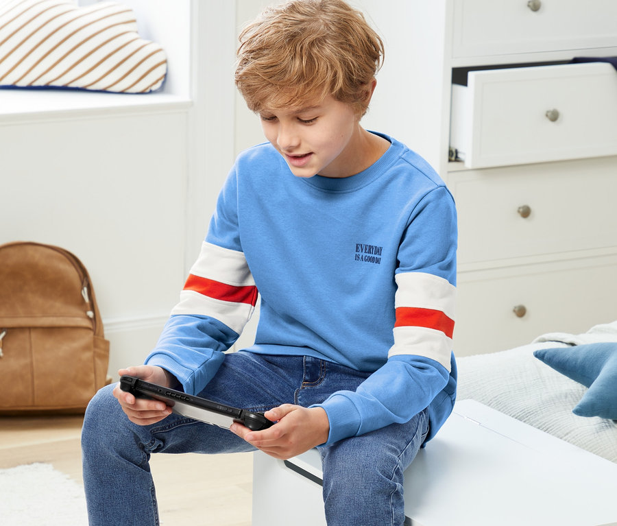 Junge sitzt und spielt ein Videospiel. Er trägt ein Sweatshirt.