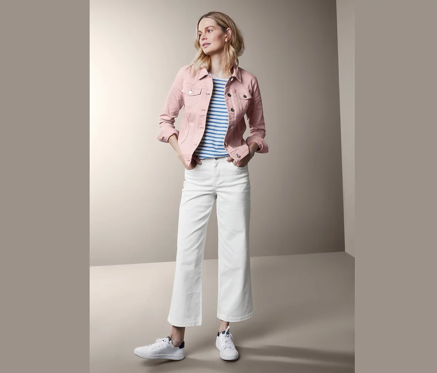 Frau posiert in roséfarbener Jeansjacke, weißer Hose und Turnschuhen.