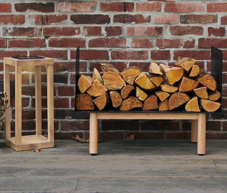 An einer Ziegelwand hängt ein Kranz, darunter steht ein Kaminholz-Regal voller Holzscheite und daneben eine Solarlaterne aus Holz.