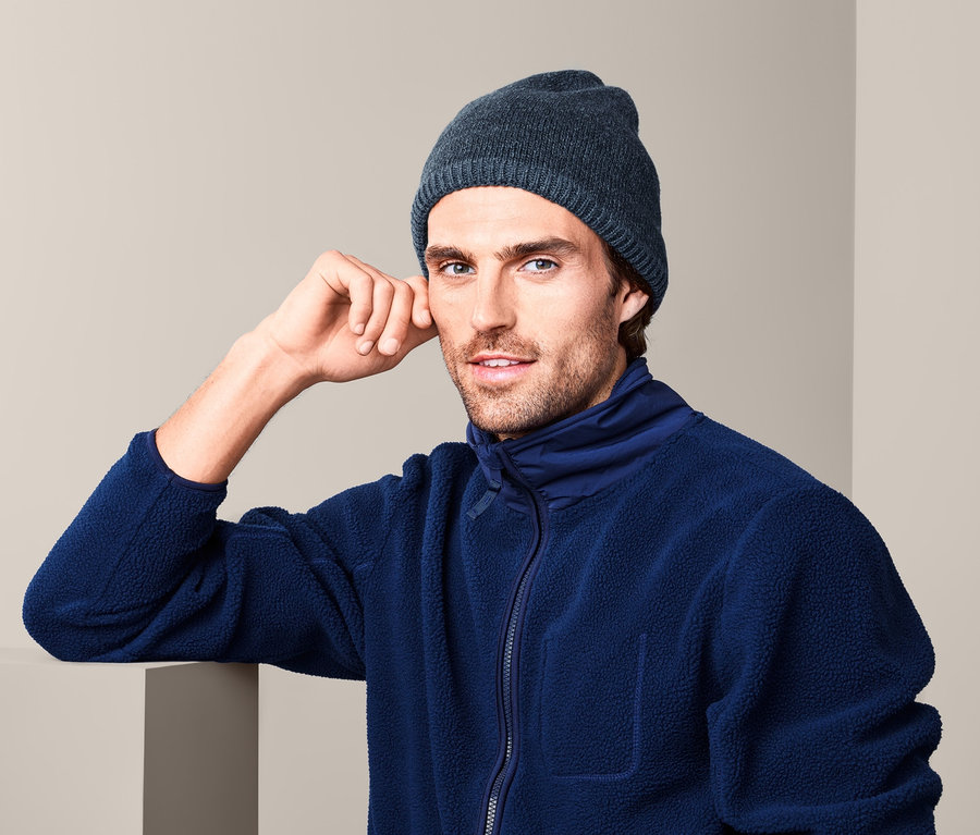 Mann mit Thermo-Strickmütze und blauer Fleecejacke lehnt an der Wand.