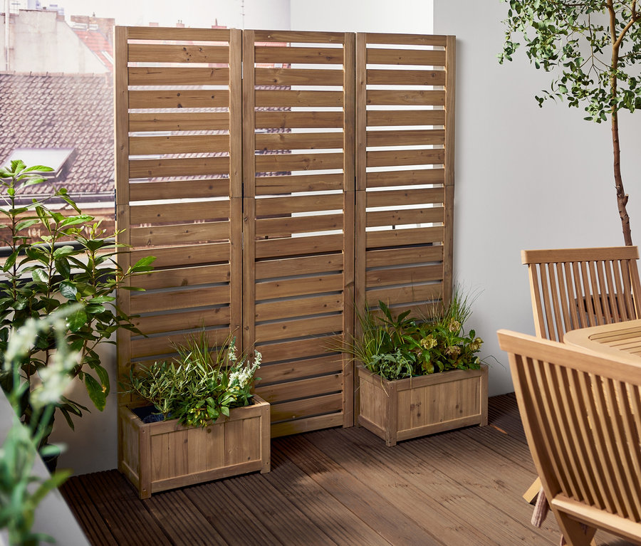 Eine Frau trägt eine Schüssel auf eine Terrasse mit einem Paravent mit Pflanztöpfen »Tinus« und einem Holzesstisch.