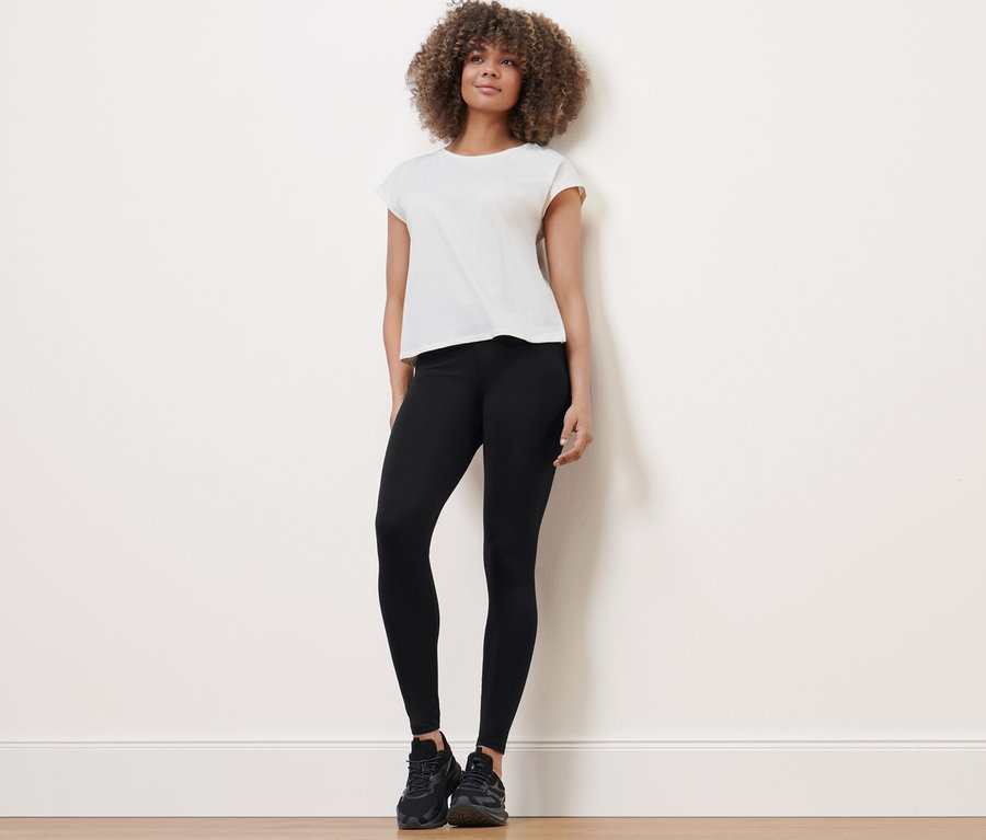 Frau steht und trägt eine Sporttight, ein T-Shirt und Turnschuhe.