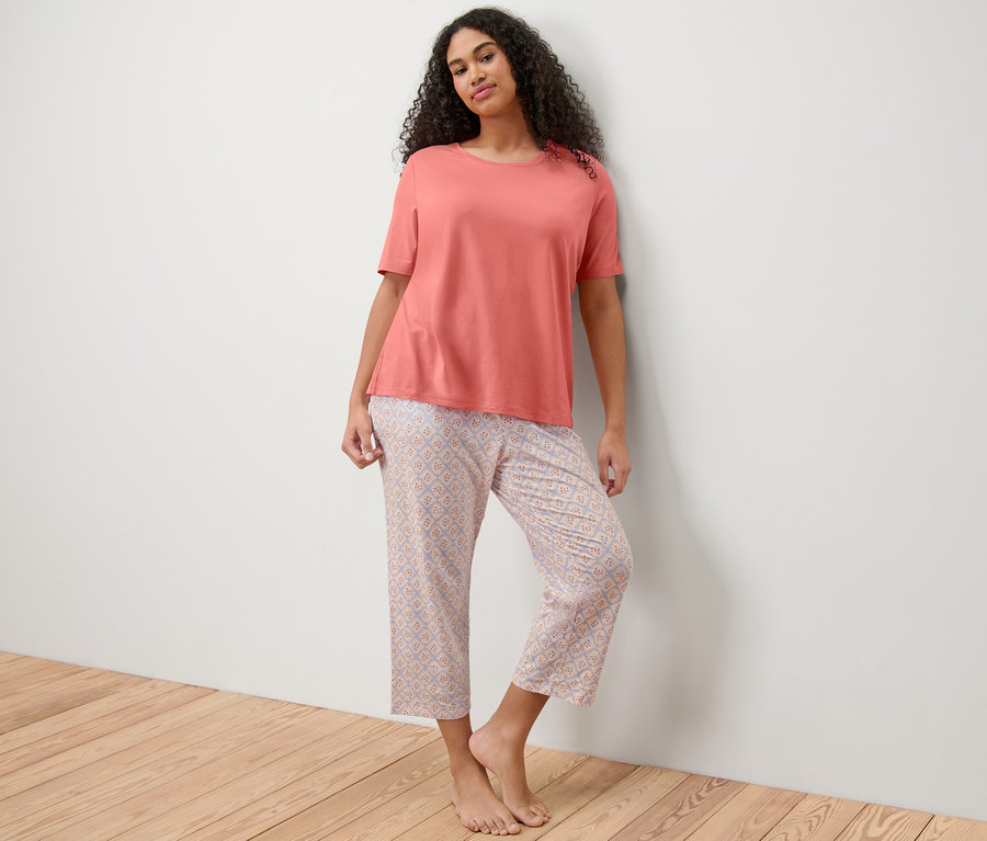 Frau lehnt an Wand und trägt lachsfarbenes Pyjama-T-Shirt und 7/8-Pyjamahose mit Muster.